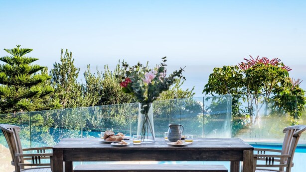 Un tavolo di legno con un vaso di fiori, una brocca e piatti di cibo, con vista su una piscina e sulla vegetazione circostante.