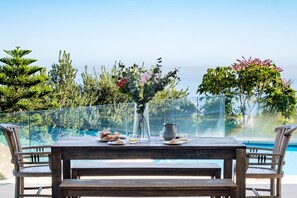Ein Holztisch mit Blumen in einer Vase, einer Karaffe und Tellern voller Speisen, mit Blick auf einen Pool und die grüne Umgebung.
