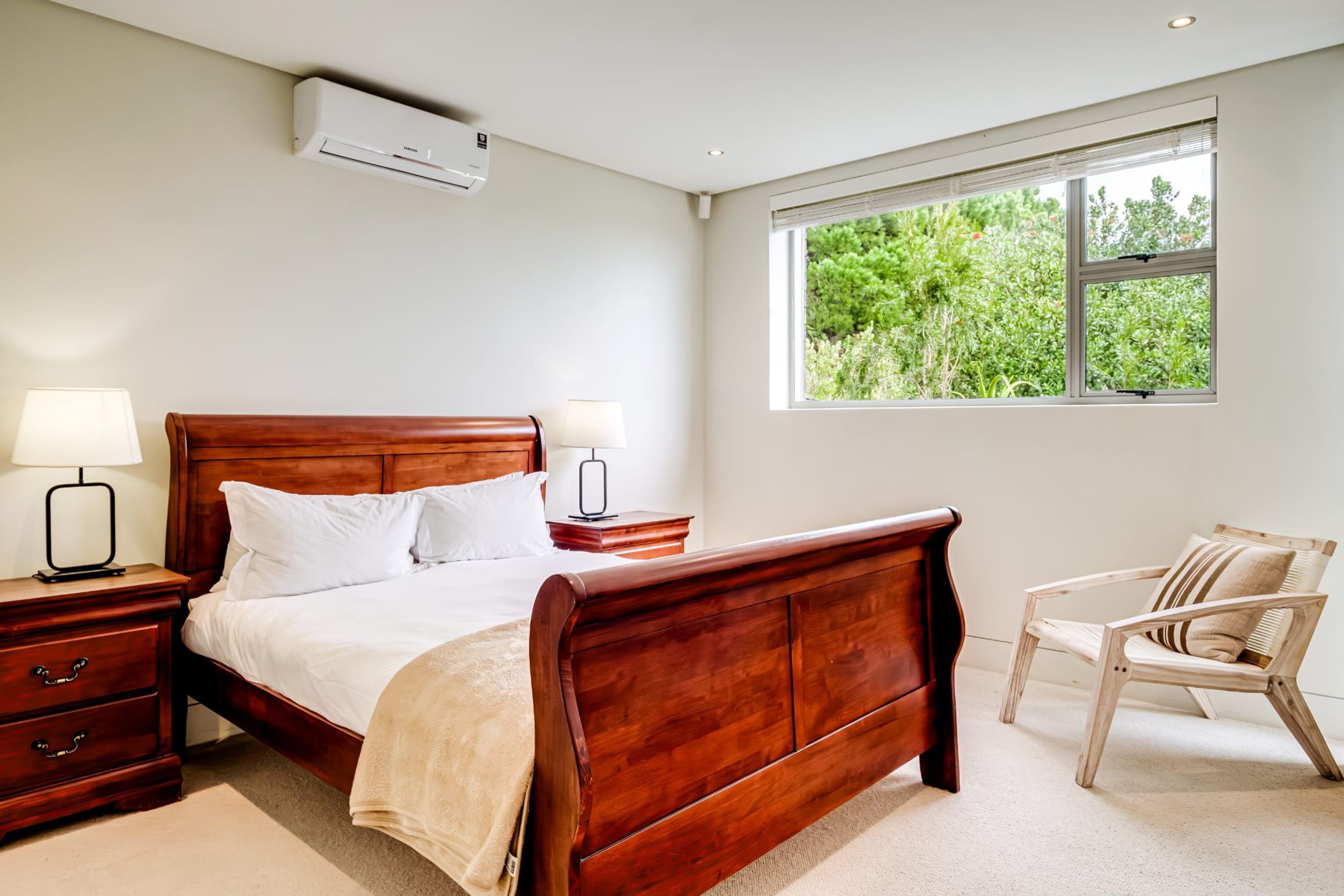 A bedroom with a wooden bed, bedside lamps, a window with a view of greenery, and a wooden chair.