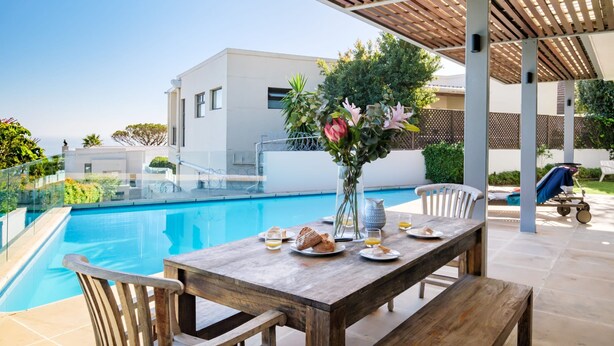 A poolside dining area with a wooden table set for two, a vase with flowers, and a view of a modern building and pool. - Sea View Sanctuary at Shakawe Villa (Cape Town)