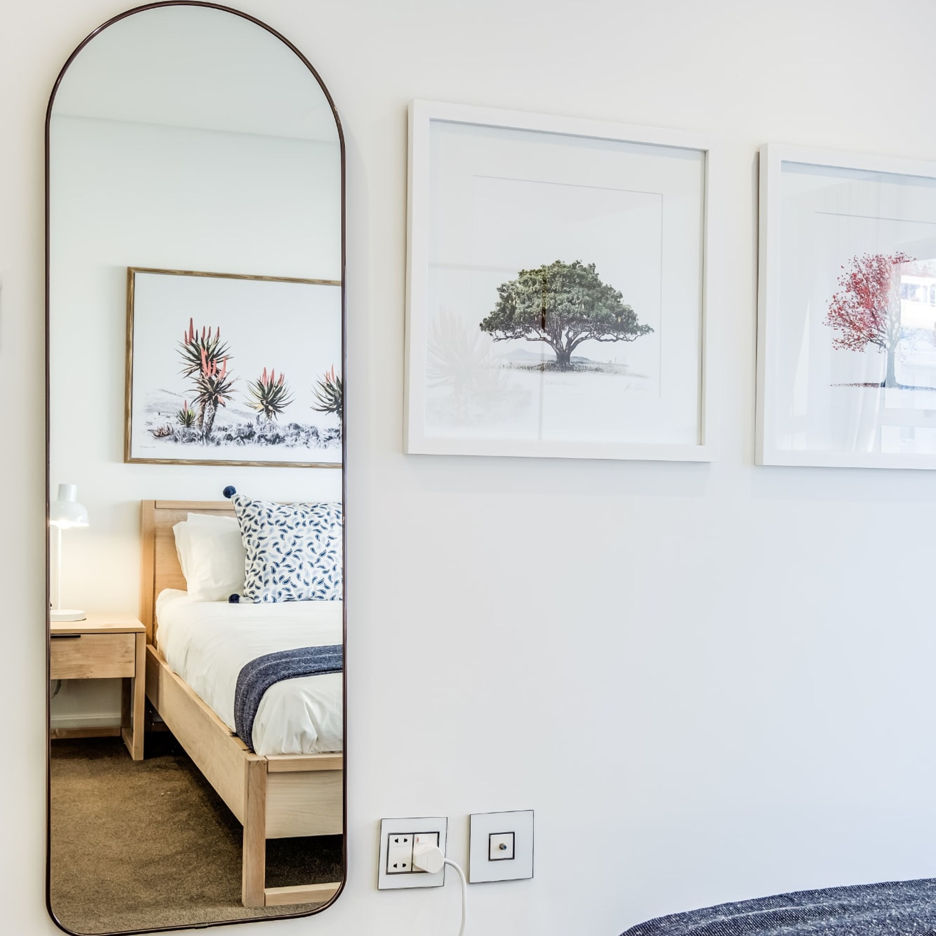 Quarto com cama, mesa de cabeceira, abajur e espelho fixado na parede. Três quadros decoram a parede acima da cama.