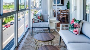 A spacious balcony with large windows, a seating area with a sofa and chairs, a small table, and a dining table with chairs.