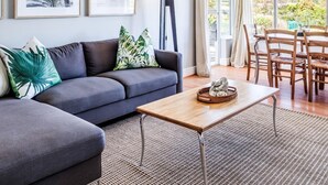 A modern living room with a grey sofa, tropical-themed cushions, a wooden coffee table, and a floor lamp. There are two framed pictures on the wall, a dining area with wooden chairs, and a large window offering a view of greenery and hills.