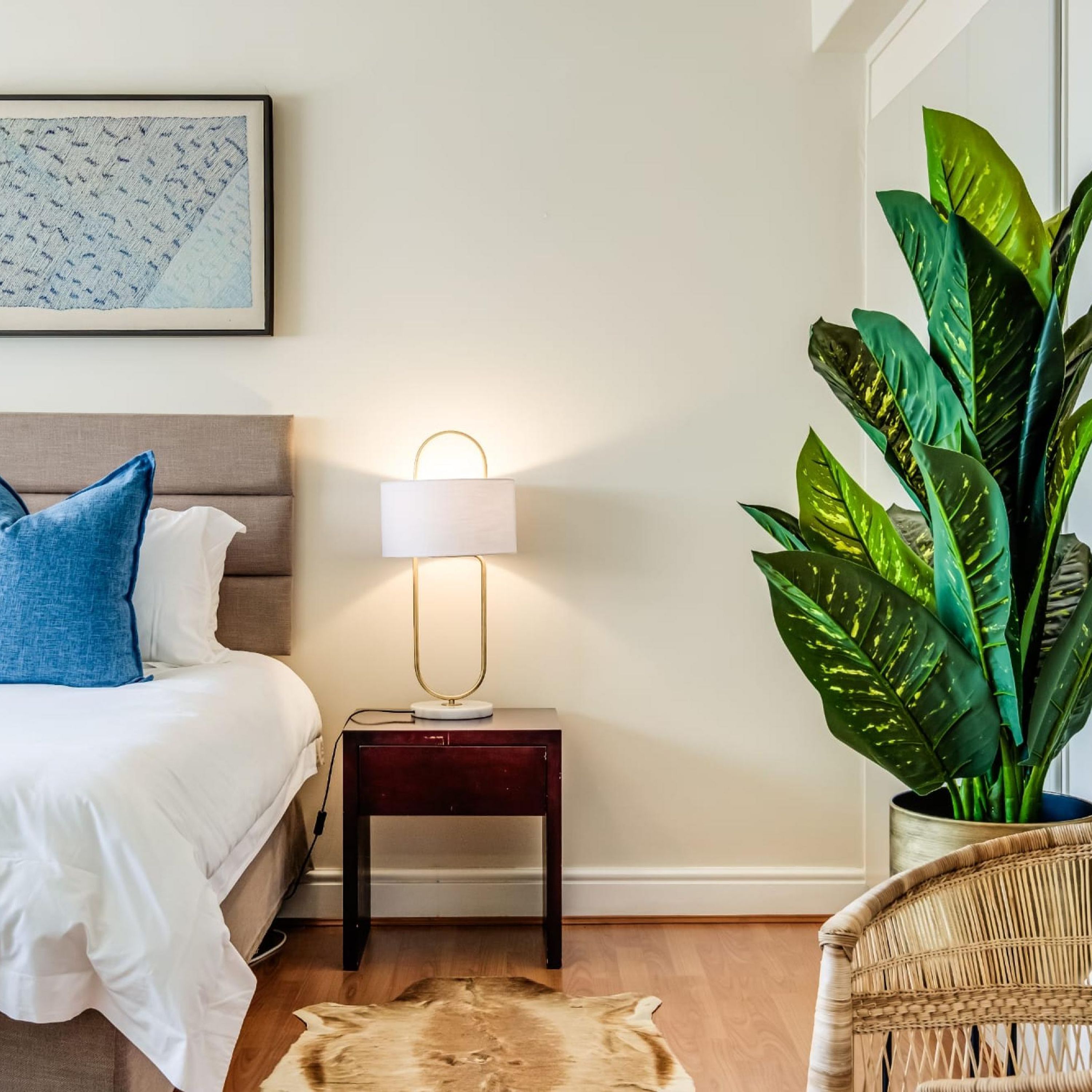 A bedroom with a bed, a nightstand with a lamp, a large potted plant, a woven chair, and a rug.