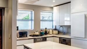 A modern kitchen with white cabinetry, a central island, and a window with blinds. - Picturesque Seaside Haven Aretzi (Cape Town)