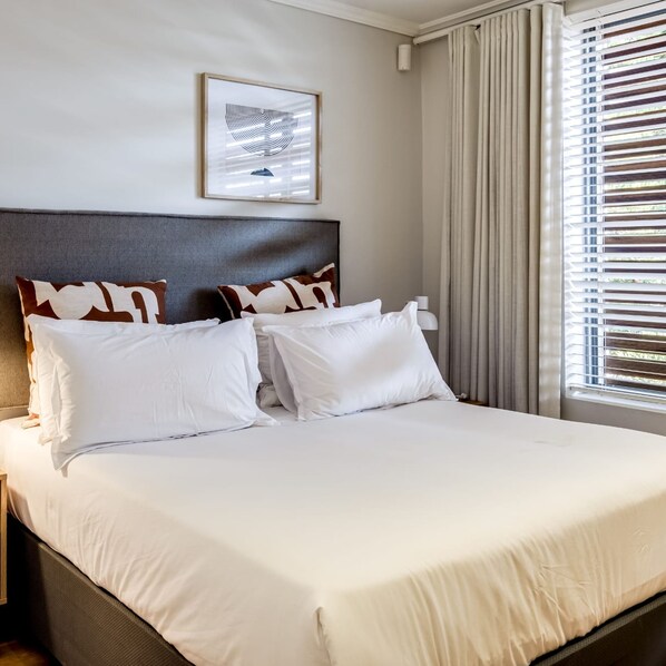 A neatly made bed with white and brown pillows, a framed picture on the wall, and a window with blinds. - Tranquil Escape in Sea Point Aureto (Cape Town)