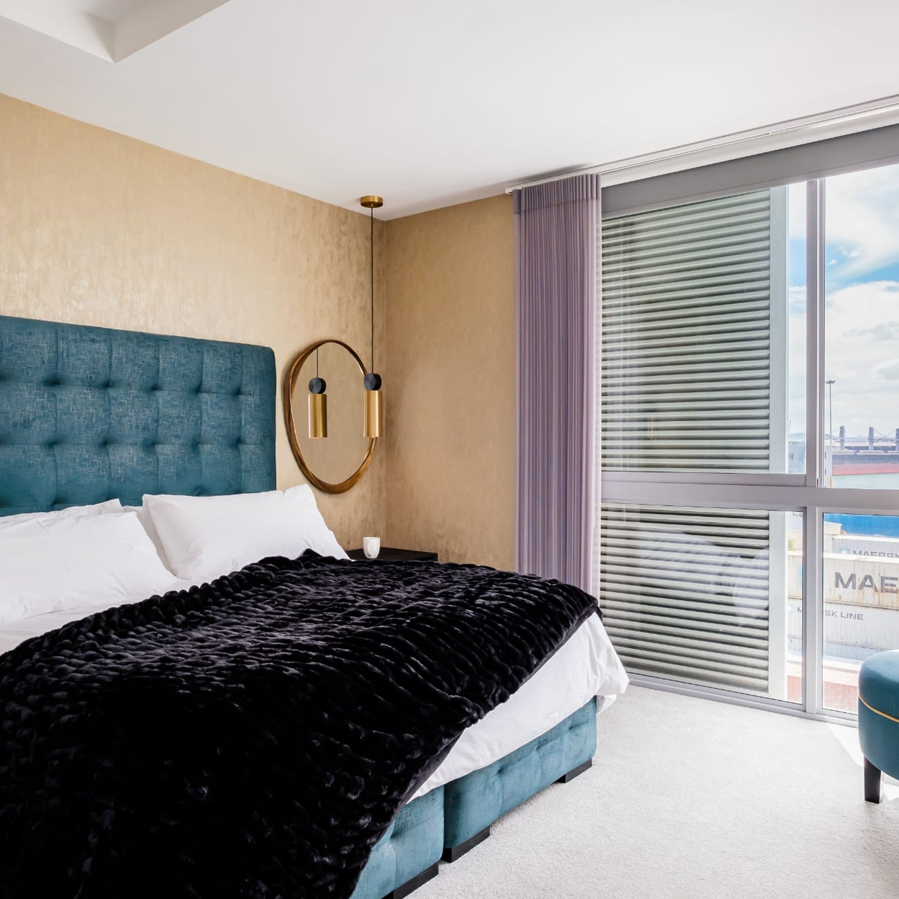 A modern hotel room with a large bed, a round wall clock, and a view of the city through large windows.