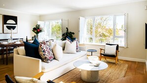 A modern living room with a sofa, coffee table, and chairs, large windows with a view of trees, and a dining area with a table and chairs. - Modern Elegance Meets City Serenity at Meliora (Cape Town)