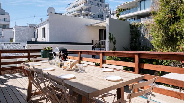 A wooden deck with a table set for four, surrounded by a wooden fence, overlooking a modern building with balconies. - Modern Elegance Meets City Serenity at Meliora (Cape Town)
