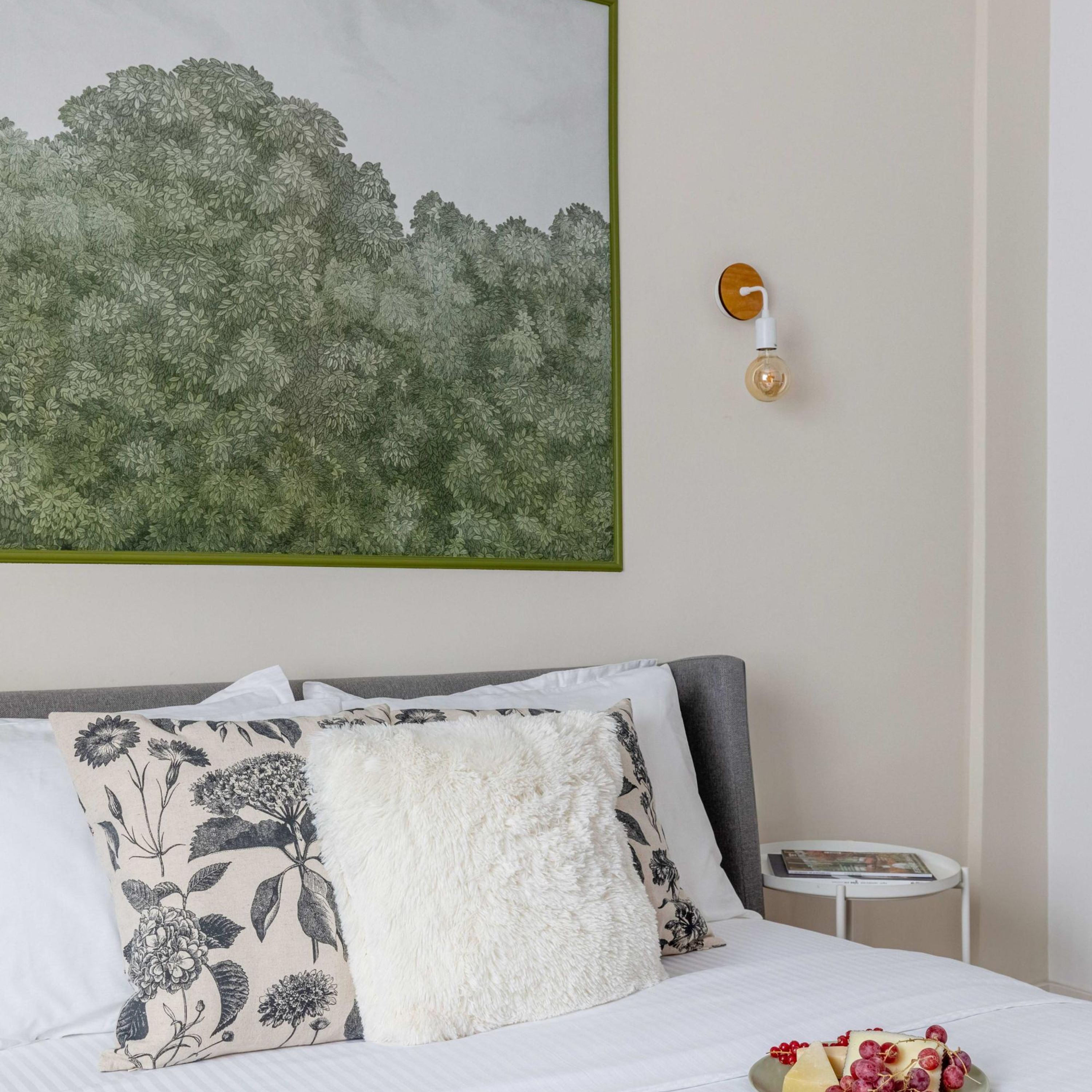 Ein Schlafzimmer mit einem Bett, Kissen, einem Nachttisch, einer Wandleuchte und einem Bilderrahmen mit einer botanischen Darstellung.