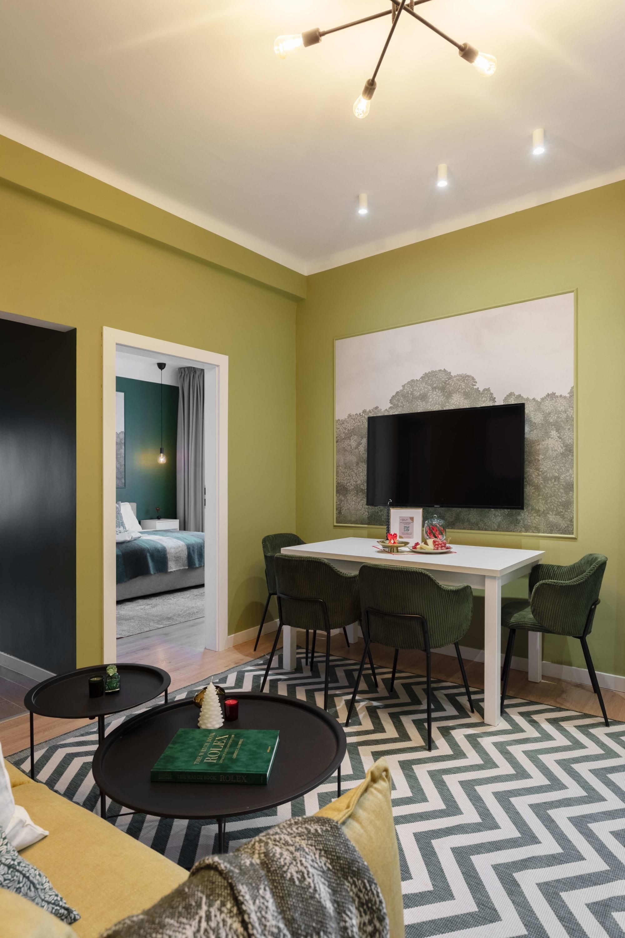 A modern hotel room with a dining area, a large flat-screen TV mounted on the wall, and a cozy seating area with a patterned rug.