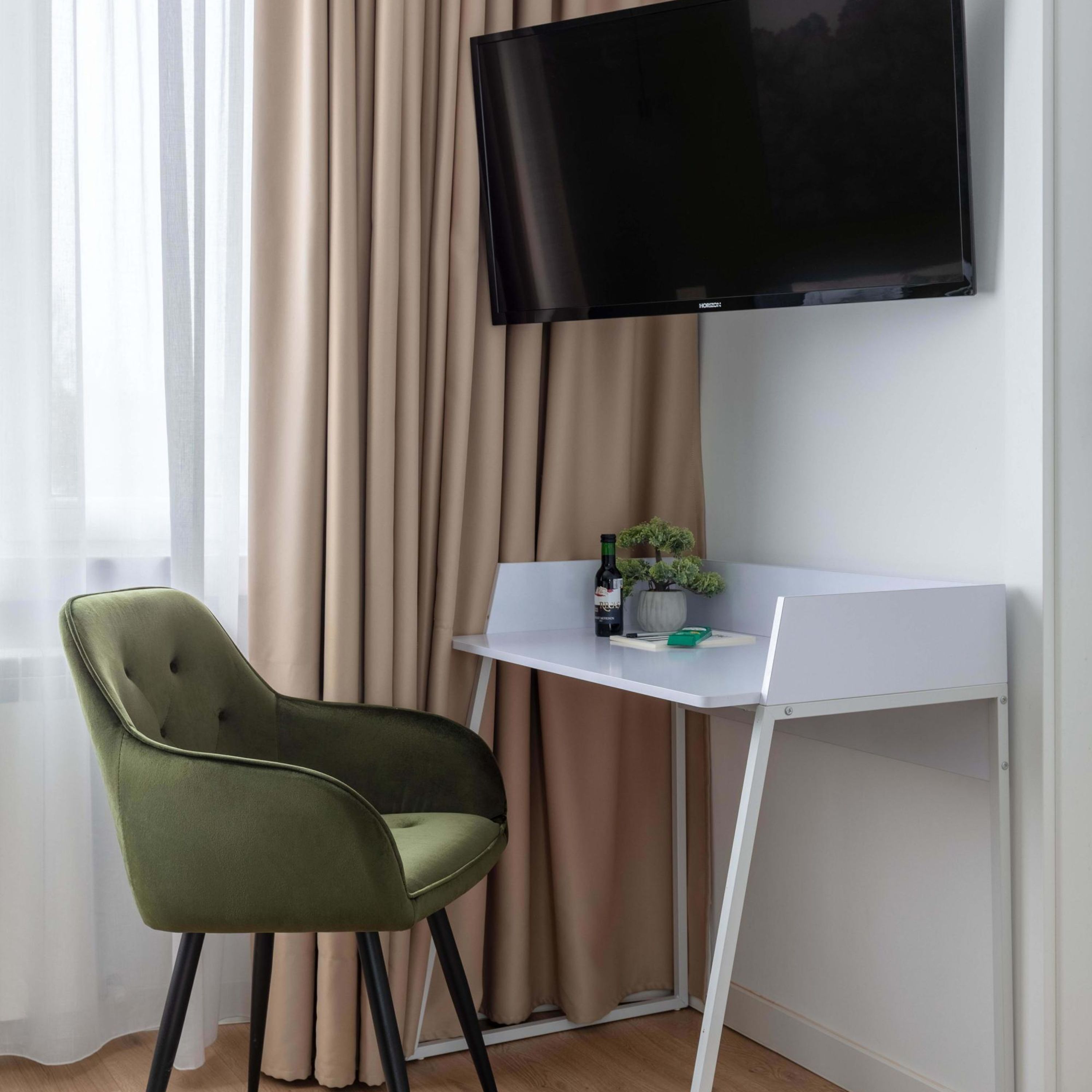 Une chambre moderne avec un fauteuil vert à revêtement en tissu, un bureau blanc, un téléviseur à écran plat fixé au mur et une fenêtre ornée de rideaux beiges.