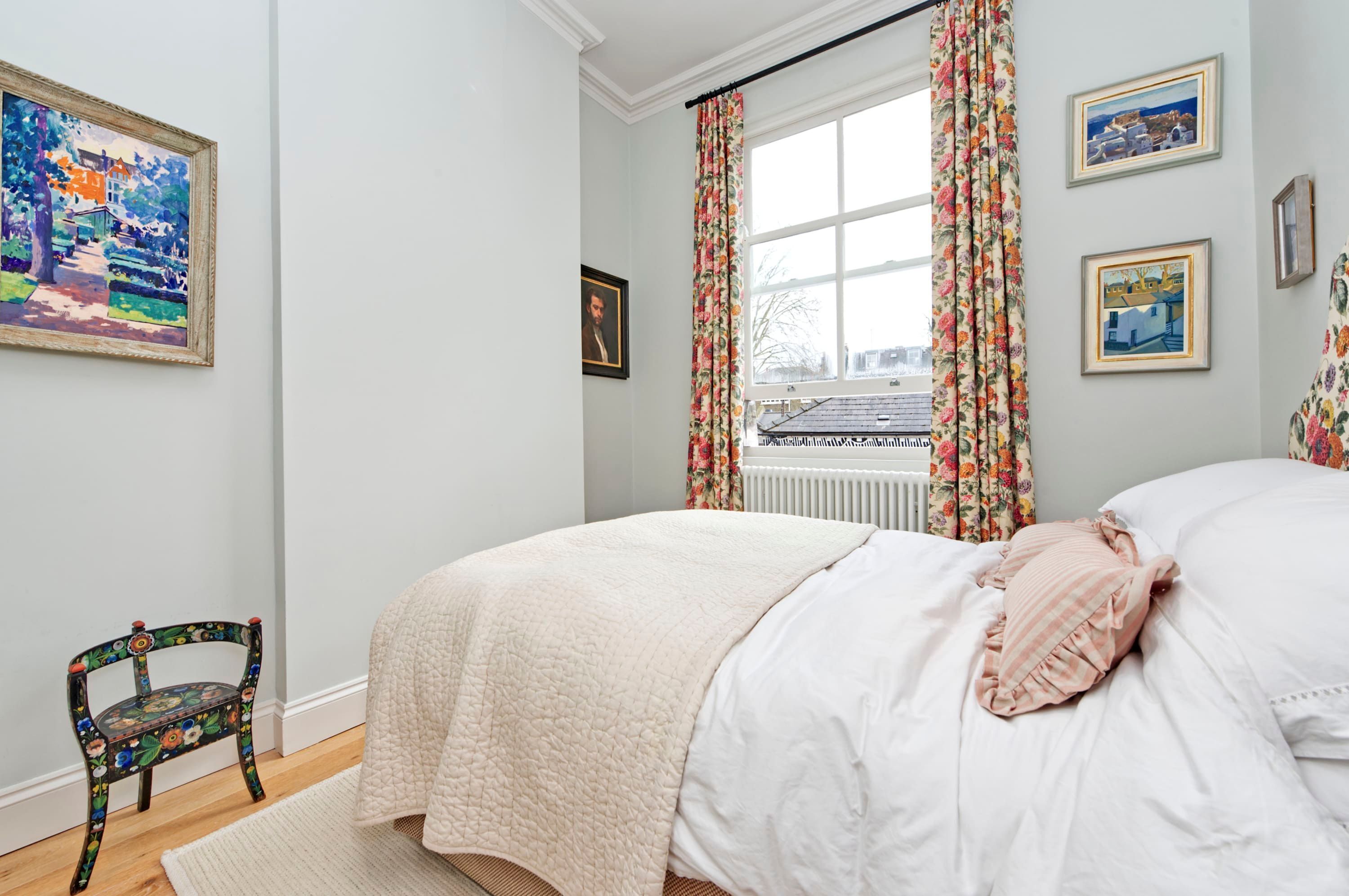 A bedroom with a bed, a chair, a window with floral curtains, and two framed pictures on the wall.