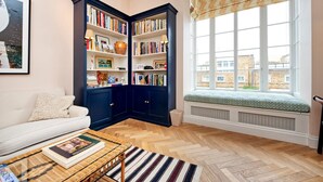 A cozy living room with a white sofa, a wooden coffee table, a dark blue bookshelf, and a window with a view of buildings. - Charming 1 Bedroom 1 Bathroom Notting Hill (London)