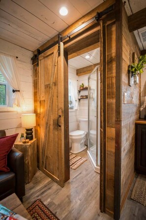 A rustic bathroom with wooden walls, a glass shower enclosure, and a toilet. - Cozy Cabin Little Red Hen 12 min to Magnolia (Waco)