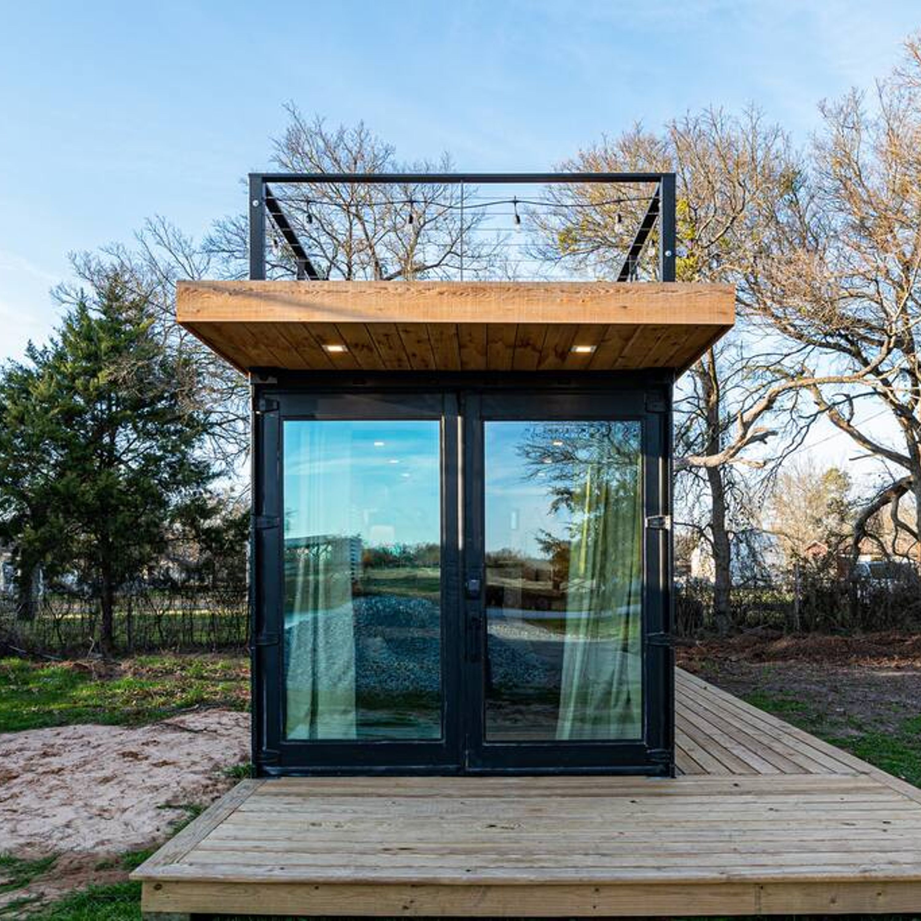 A modern, minimalist structure with a wooden deck and large glass doors, surrounded by trees.