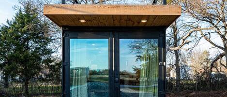 A modern, minimalist structure with a wooden deck and large glass doors, surrounded by trees.
