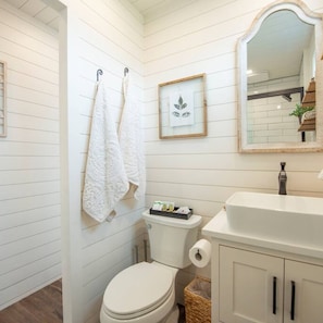 A bathroom with white wooden walls, a toilet, a sink with a mirror, and two white towels hanging on hooks.