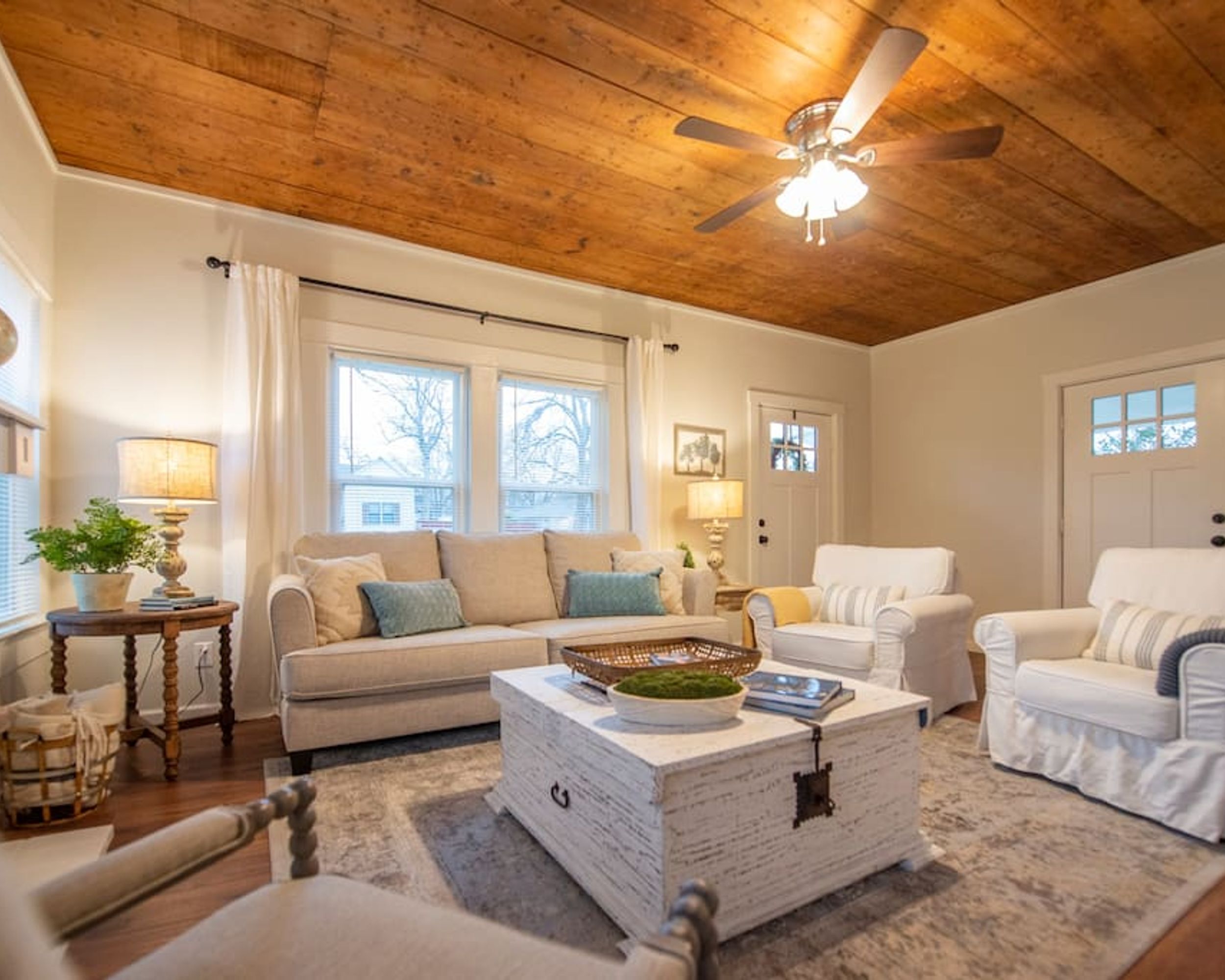 Una sala de estar acogedora con techo de madera, un sofá blanco, un sillón blanco y una mesa de centro con objetos decorativos.