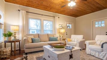 Una sala de estar acogedora con techo de madera, un sofá blanco, un sillón blanco y una mesa de centro con objetos decorativos.