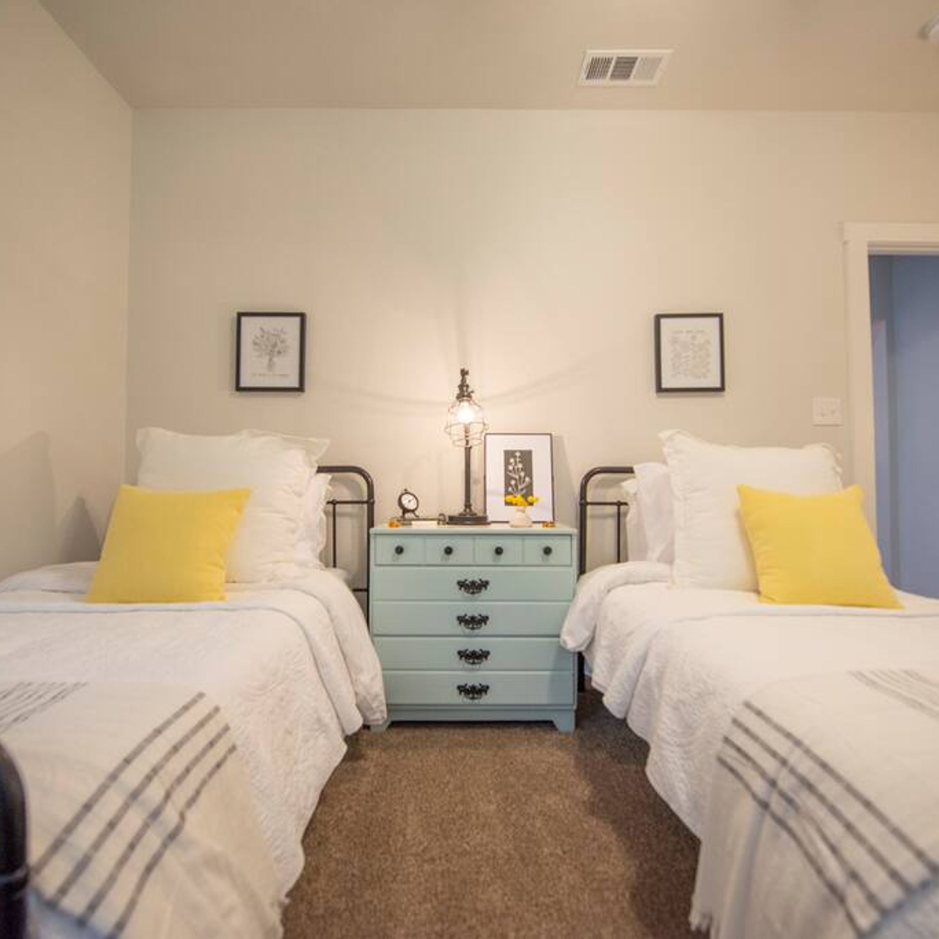 A bedroom with two beds, each with white bedding and yellow pillows, a light blue dresser with drawers, and framed pictures on the walls.