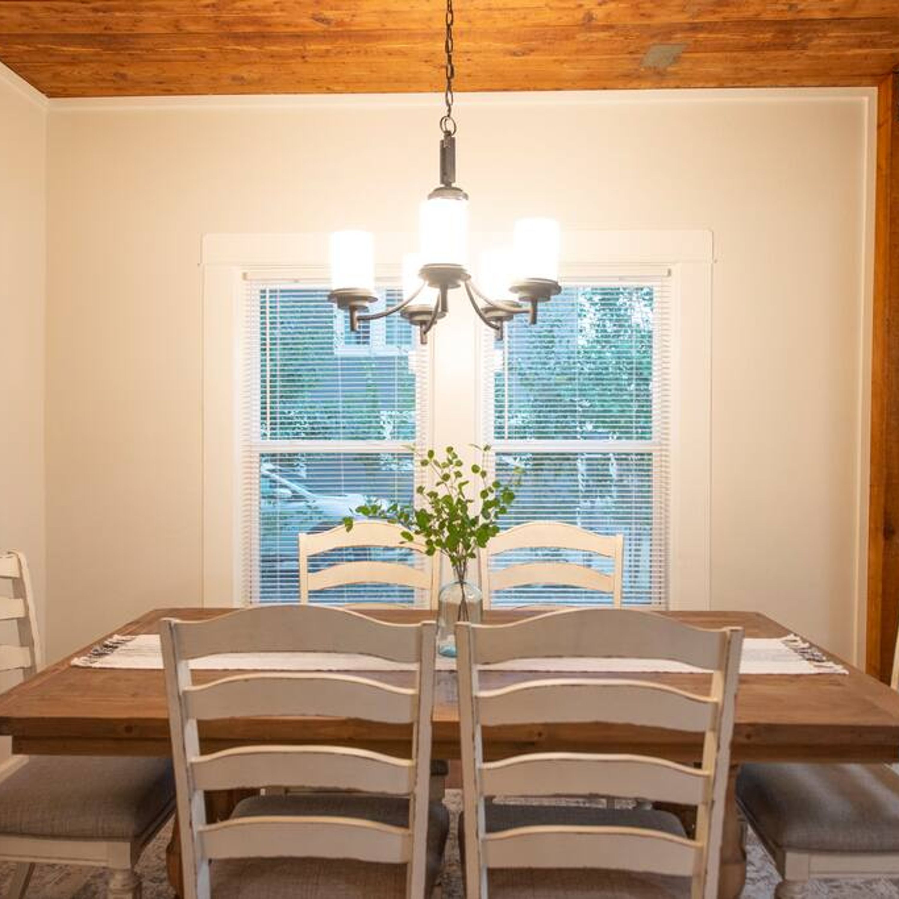 A dining area with a wooden table, four chairs, a chandelier, and a window with blinds.