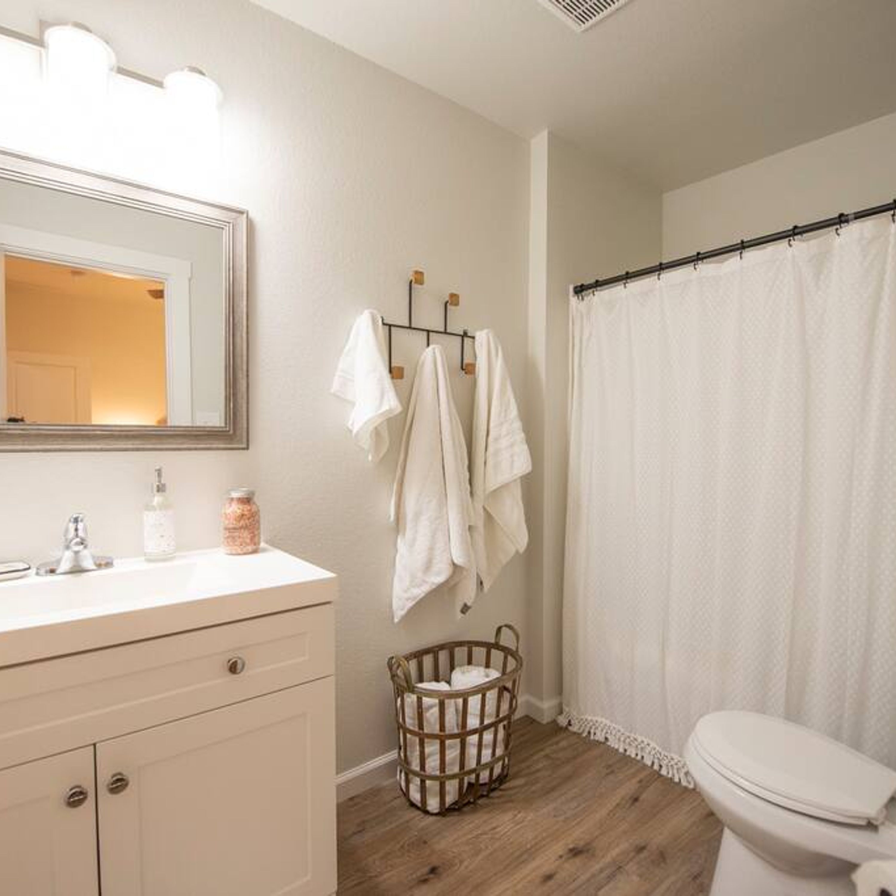 Un baño con suelo de madera, inodoro blanco, lavabo con espejo y cortina de ducha.