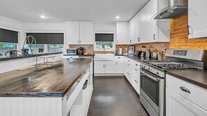 Eine moderne Küche mit weißen Schränken, einer Holz Arbeitsplatte, Edelstahlgeräten und Blick ins Freie durch die Fenster.