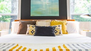 Ein Schlafzimmer mit einem großen Bett, mehreren Kissen, einem Bild an der Wand und Fenstern mit Blick auf Grünflächen.