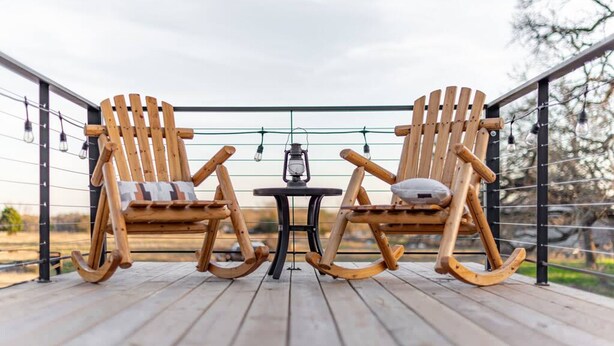 Two wooden rocking chairs on a wooden deck with a table in between, under a string of lights. - Deer Creek Tiny Container Home-12 Min to Magnolia (Waco)