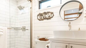 A bathroom with a glass shower enclosure, a white toilet, a sink with a mirror above it, and a decorative wall piece. - Deer Creek Tiny Container Home-12 Min to Magnolia (Waco)