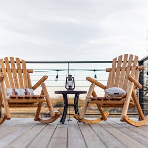 Deux fauteuils à bascule en bois, placés face à face et dotés de coussins, une petite table avec une lanterne, et une vue sur le ciel.