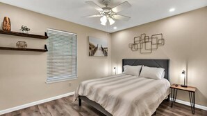A bedroom with a bed, wooden flooring, a ceiling fan, wall-mounted shelves, a window with blinds, and a wall art piece.