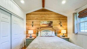 A bedroom with a wooden ceiling, a bed with a beige blanket, two bedside tables with lamps, and a window with curtains. - Wagon Wheel Cottage Couples Getaway in the Country (Waco)