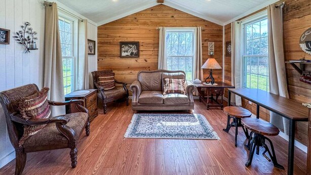 A cozy living room with wooden walls, a large window, a fireplace, and a dining area. - Wagon Wheel Cottage Couples Getaway in the Country (Waco)
