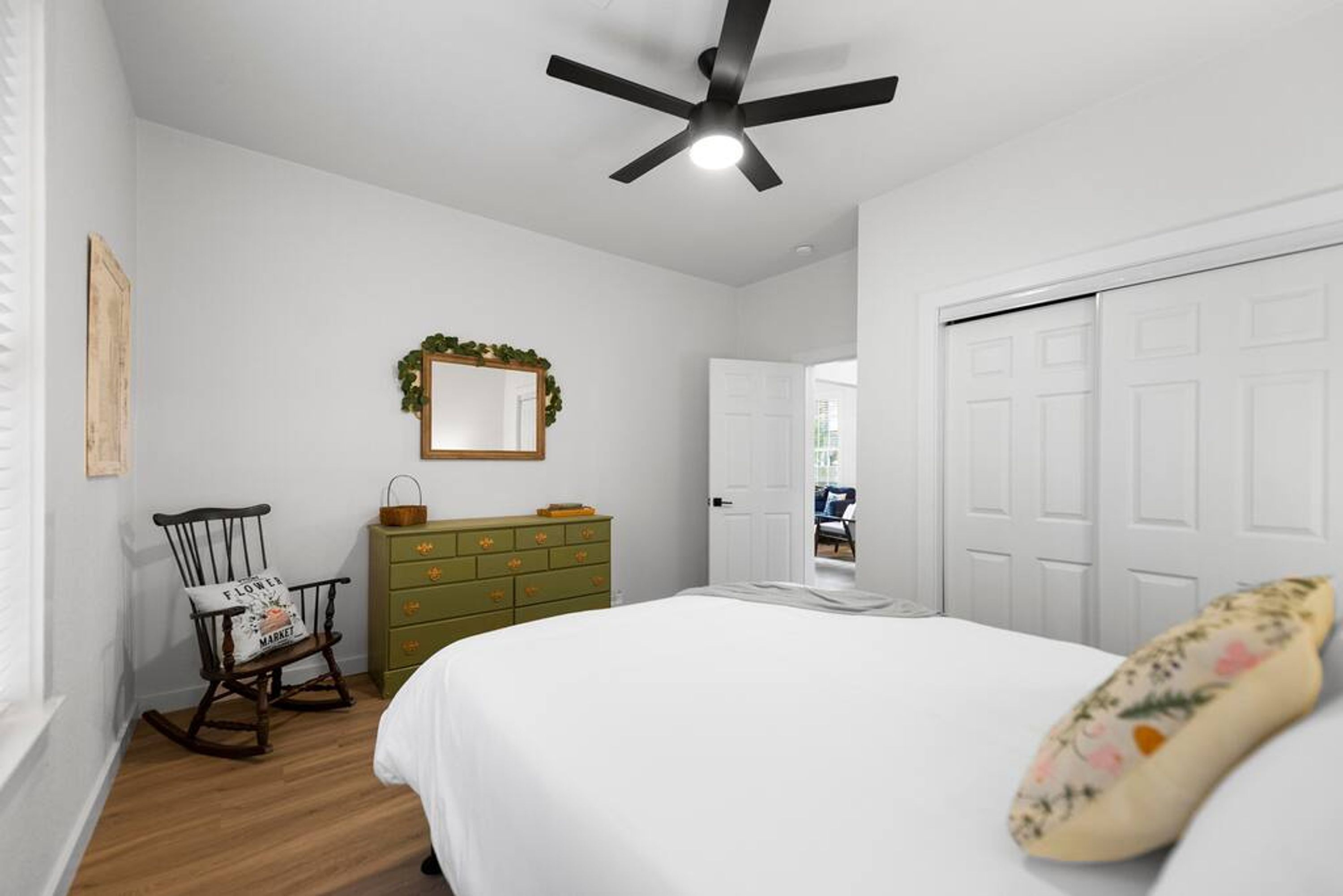 A bedroom with a bed, a rocking chair, a dresser, a ceiling fan, and a door.