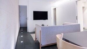 Een moderne woonkamer met een flatscreen televisie aan de muur, een grijze bank, een houten eettafel en een klein vloerkleed.