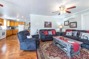 A spacious living room with a dark grey sofa set, a wooden coffee table, and a red rug. A kitchen is visible in the background with wooden cabinets and a white refrigerator. - Songbird Haven Cozy Country Getaway (Waco)