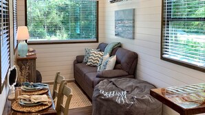 A cozy living room with a brown sofa, a wooden coffee table, and a bean bag chair. There are two windows with blinds, a painting on the wall, and a ceiling with exposed beams. - The Shoreline Container Tiny Home 12 min to Magnolia Baylor (Waco)