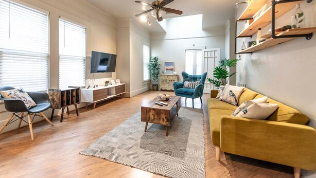 A spacious living room with a wooden floor, a ceiling fan, a TV, a yellow sofa, a blue chair, and a coffee table. - New Gathering Place on Columbus (Waco)