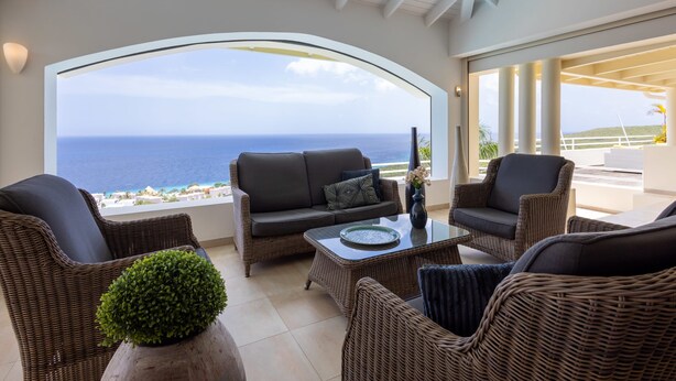 A spacious balcony with wicker furniture, a glass table, and a view of the ocean. - Shoreline Harmony at Villa Solana (Sint Willibrordus)