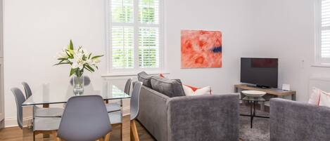 A modern living room with a dining area, grey sofas, a TV, and a large window with blinds.