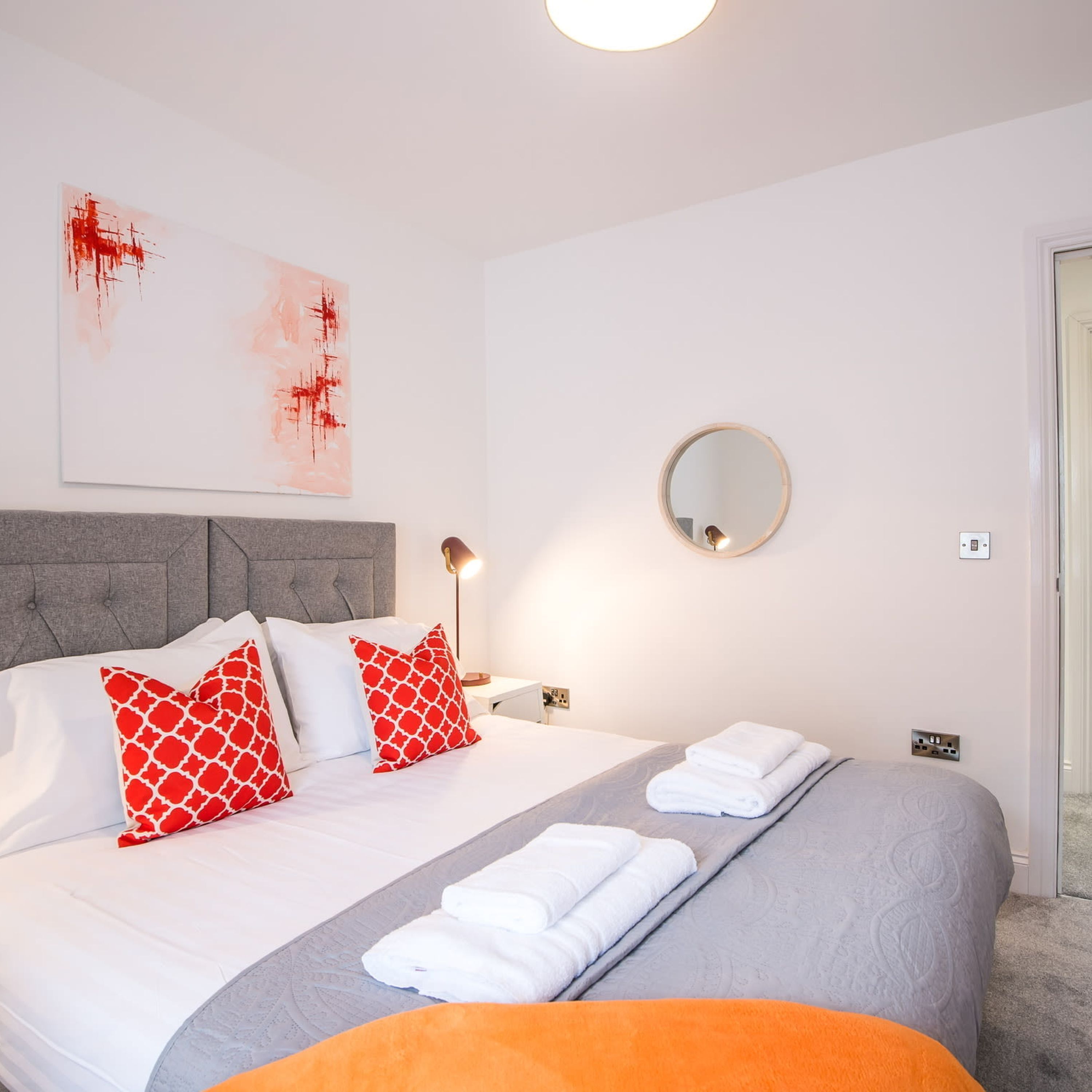 Een netjes opgemaakt bed met kussens in wit en rood en een deken in grijs en wit. Aan de muur hangt een ronde spiegel, er staat een bedlampje en er ligt een opgevouwen witte handdoek op het bed.