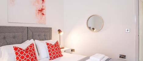 A neatly made bed with white and red patterned pillows, a grey headboard, and a grey and white patterned bedspread. There is a round mirror on the wall, a bedside lamp, and a folded white towel on the bed.