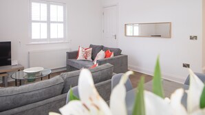 Ett modernt vardagsrum med en grå soffa, ett soffbord i glas och en platt-TV. Det finns ett fönster med persienner, ett element och en spegel på väggen.