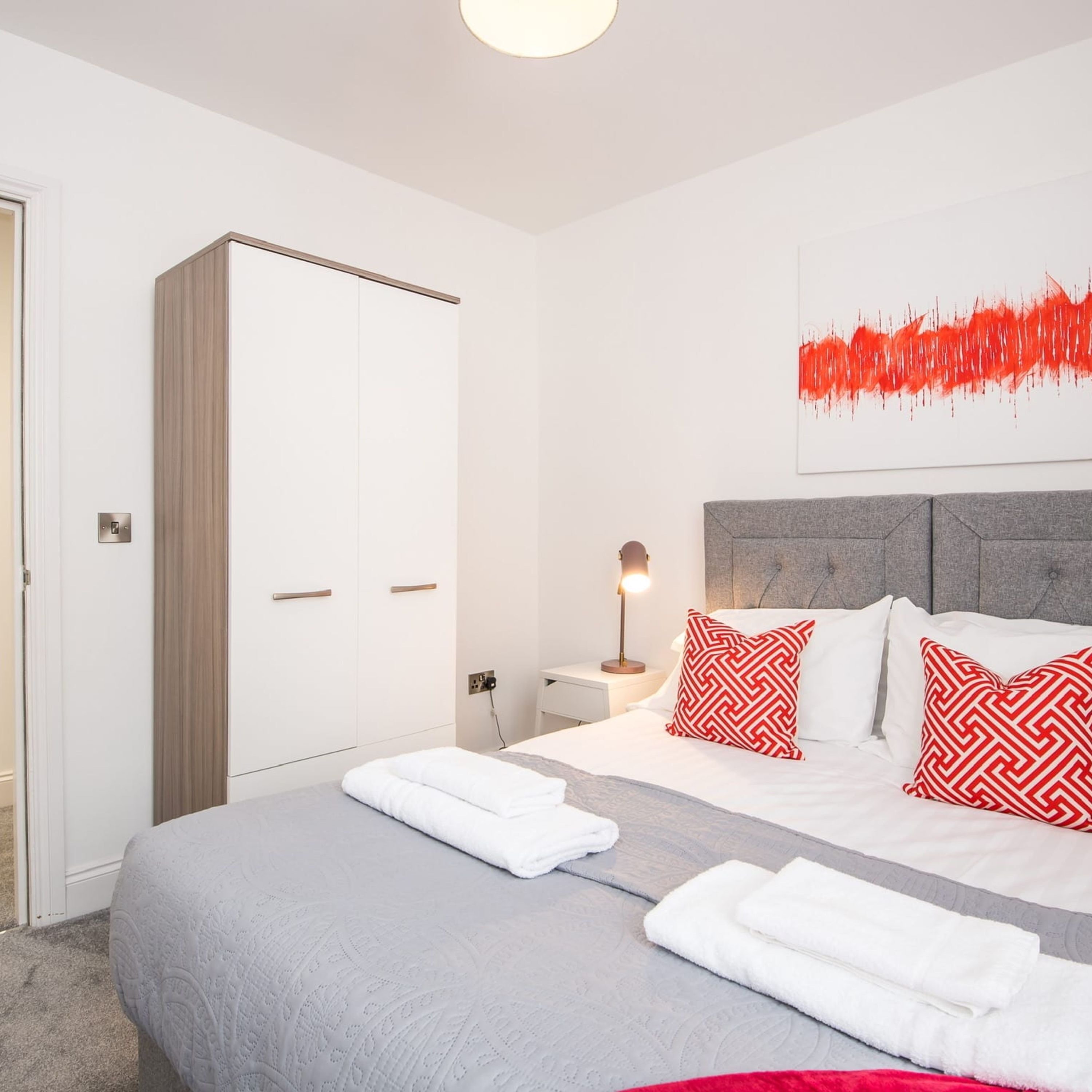 A modern bedroom with a bed, a wardrobe, a bedside table with a lamp, and a red abstract painting on the wall.