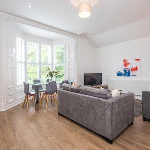 Ein modernes Wohnzimmer mit grauem Sofa, einem Esstisch mit Stühlen, einem Fernseher und einem Fenster mit Jalousien.
