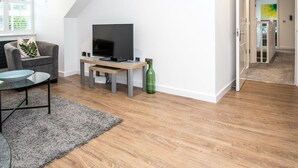 Ein modernes Wohnzimmer mit einem Flachbildfernseher auf einem Holzboden, einem grauen Sofa und einem Glastisch.