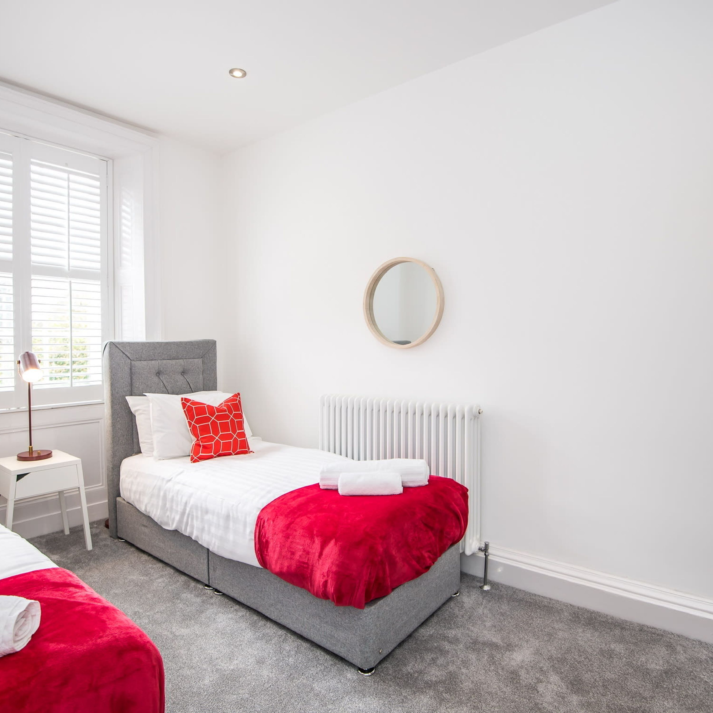 A bedroom with a grey bed, white bedding, a red throw, a small bedside table with a lamp, and a radiator.