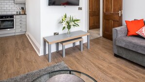 Ein modernes Wohnzimmer mit grauem Sofa, einem Flachbildfernseher an der Wand, einer Holztür und einer Küchenzeile mit grauen Schränken und einem eingebauten Backofen.